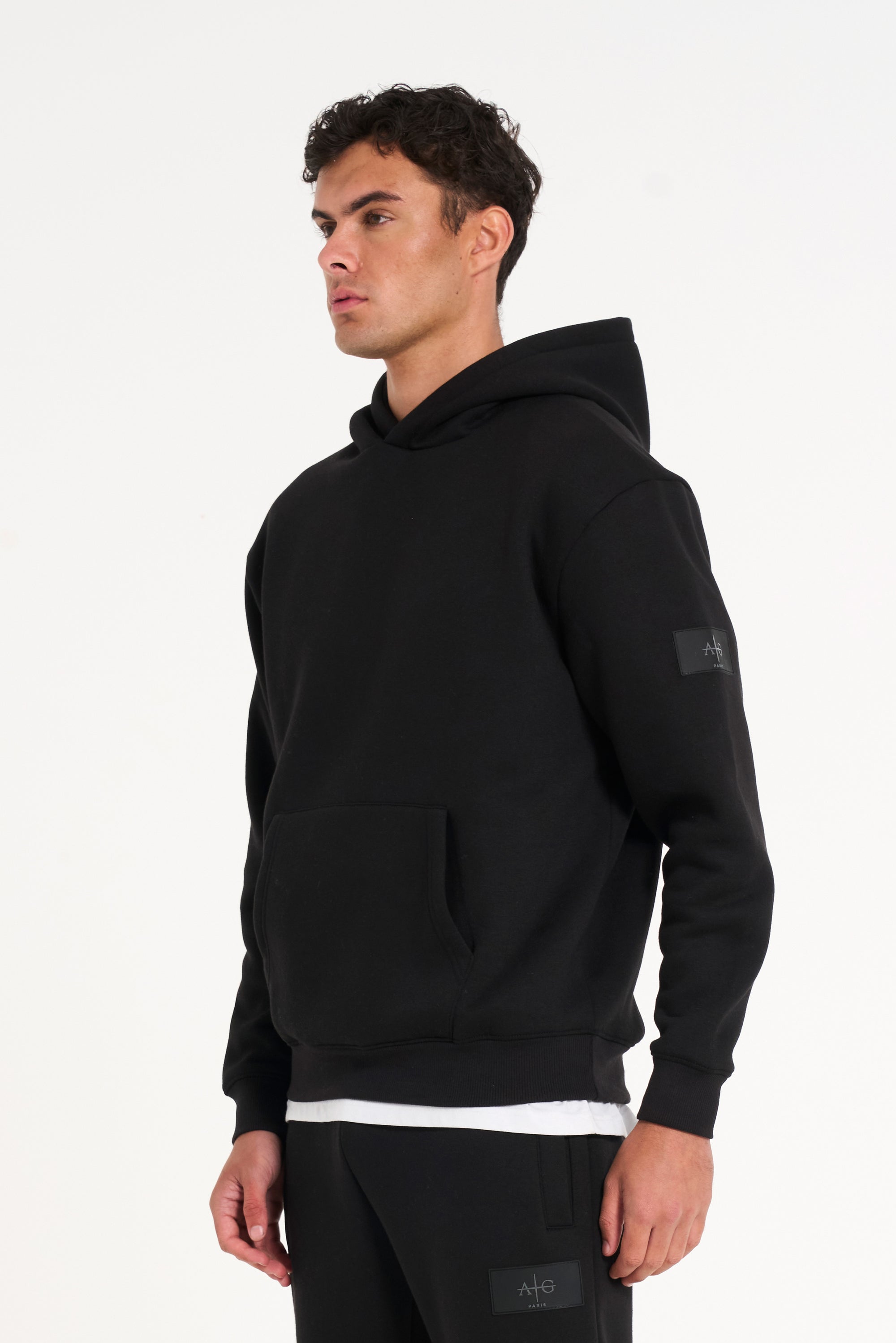 Model facing to the left wearing black tracksuit top and bottoms with badge on left arm